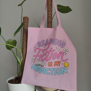 Może przedstawiać: Różowa torba tote z napisem "Reading Fiction is my Addiction" w kolorowych literach z grafiką książki i uśmiechniętej buźki.