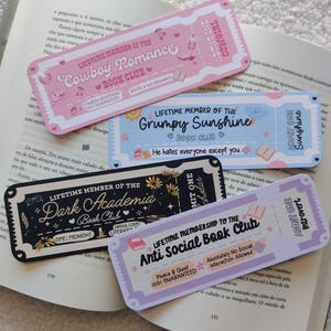 Op de afbeelding: Vier decoratieve bladwijzers met ticketontwerpen, elk met een thema voor een boekenclub. De bladwijzers bevatten teksten als "Cowboy Romance", "Grumpy Sunshine", "Dark Academia" en "Anti Social Book Club". Ze zijn in roze, blauw, zwart en lavendel.