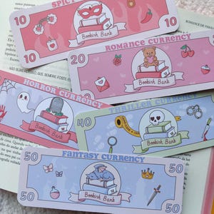 Peut inclure: Plusieurs billets de banque décoratifs avec des thèmes variés. Le "Spicy Currency" présente un masque et un piment, le "Romance Currency" un ours en peluche, le "Horror Currency" un fantôme, le "Thriller Currency" un crâne et le "Fantasy Currency" un dragon.