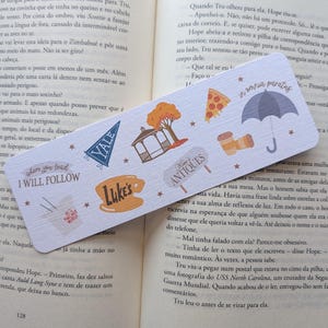 Op de afbeelding: Een rechthoekige boekenlegger met diverse illustraties en tekst, waaronder een pizzapunt, een paraplu en de woorden "Where I Will Follow" en "Luke's". De boekenlegger ligt op een open boek.