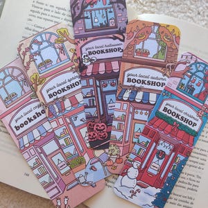 Könnte beinhalten: Vier rechteckige Lesezeichen mit illustrierten Ladenfronten, beschriftet mit your local cozy bookshop, spring, halloween, autumn und christmas bookshop, die jeweils mit saisonalen Details und Farben gestaltet sind.