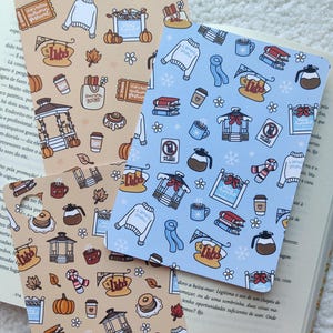 Op de afbeelding: Drie decoratieve bladwijzers met een herhalend patroon van gezellige herfst- en winterthema-illustraties. De bladwijzers bevatten pompoenen, truien, koffiekopjes en de tekst "Luke's". Eén bladwijzer is bruin en de andere lichtblauw.