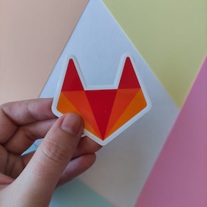 Peut inclure: Un autocollant repr&eacute;sentant une t&ecirc;te de renard rouge et orange avec un contour blanc. L'autocollant est tenu par une main sur un fond de couleurs pastel.