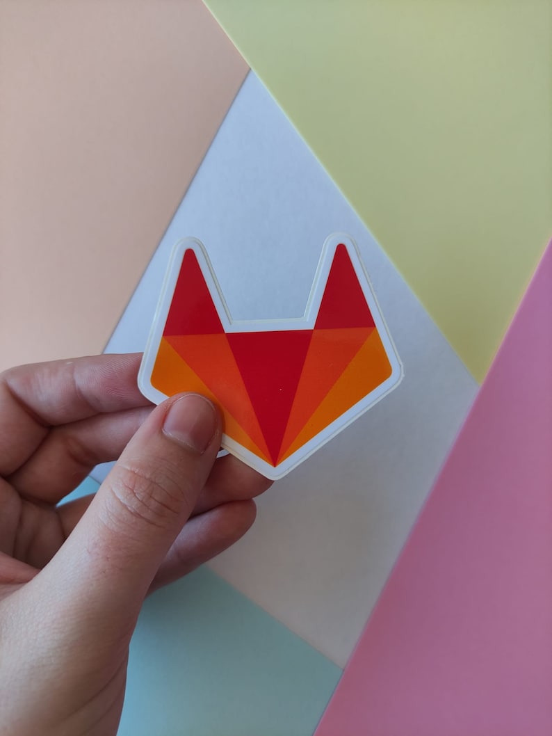 Peut inclure: Un autocollant repr&eacute;sentant une t&ecirc;te de renard rouge et orange avec un contour blanc. L'autocollant est tenu par une main sur un fond de couleurs pastel.