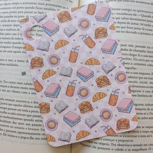 Op de afbeelding: Een hartvormige bladwijzer met een herhalend patroon van ontbijtproducten: pannenkoeken, croissants, koffie, boeken en ijsdranken. De achtergrond is een lichtroze geruit ontwerp. De bladwijzer ligt op een open boek.