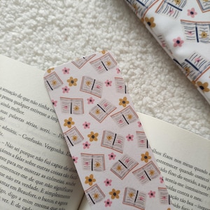 Peut inclure: Un marque-page blanc avec un motif répété de fleurs roses et jaunes et de livres ouverts. Le marque-page est posé sur un livre avec du texte en portugais.