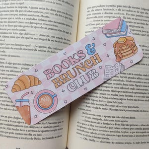 Op de afbeelding: Een bladwijzer met de tekst "BOOKS & BRUNCH CLUB" in een retro lettertype, met illustraties van pannenkoeken, koffie en een croissant. De bladwijzer ligt op een open boek met tekst.