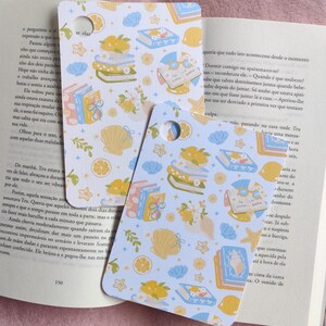 Op de afbeelding: Twee decoratieve bladwijzers met een lichtblauwe achtergrond. De bladwijzers bevatten illustraties van boeken, citroenen, schelpen en bloemen. De tekst "se elas" is zichtbaar op een bladwijzer. De bladwijzers liggen op een open boek.