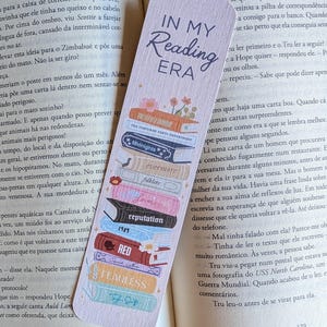 Peut inclure: Un marque-page en papier rectangulaire avec un fond violet clair portant le texte IN MY READING ERA en haut. Sous le texte se trouve une pile verticale de livres illustrés colorés avec divers titres, dont Midnights, folklore et RED.