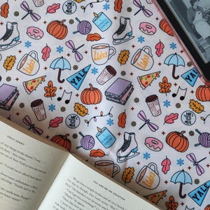 May include: A white fabric featuring a repeating pattern of autumn-themed illustrations, such as pumpkins, leaves, coffee cups, and ice skates. A book and an e-reader are visible, suggesting a cosy reading theme.