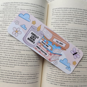 Pode incluir: Um marcador de livros colorido com o texto "Smells Like New Books" e "I'll Just Read One More Chapter". O marcador apresenta ilustrações de livros, nuvens, flores e uma vela. Está colocado num livro aberto.