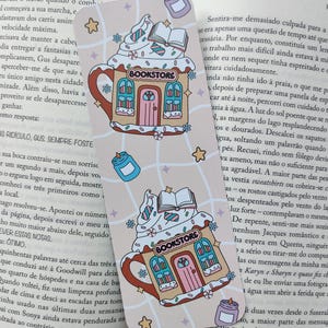 Pode incluir: Um marcador de livros caprichoso com um design de livraria em forma de caneca de café com chantilly e um livro em cima. O marcador de livros é rosa claro com detalhes em azul, rosa e castanho. Inclui a palavra "Bookstore".