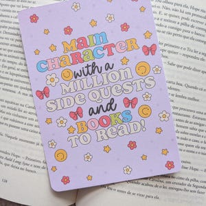 Puede incluir: Un marcapáginas lavanda con el texto "Main Character with a Million Side Quests and Books to Read!" en letras coloridas y divertidas. El marcapáginas está decorado con flores, estrellas y lazos.