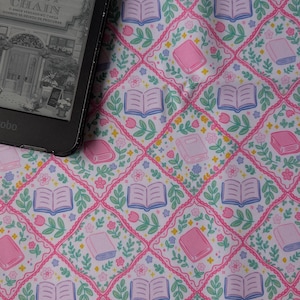 Op de afbeelding: Een zwarte e-reader met de boekomslag A Florista Daisy Chain, geplaatst op een witte stof met een herhalend ruitpatroon. Het patroon bevat illustraties van open en gesloten boeken, roze tulpen en kleine paarse bloemetjes.