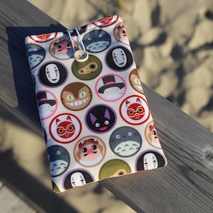 Op de afbeelding: Een witte telefoonhoes van stof met een kleurrijk patroon van cartoonfiguren uit de film Spirited Away. De personages zijn onder andere No Face, Totoro, Jiji en anderen. De hoes heeft een witte knop en een wit touwtje.