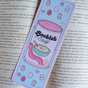 Pode incluir: Um marcador de página retangular com cantos arredondados, fundo azul claro e uma ilustração de uma lata de sopa rosa. A lata está rotulada como Bookish Soup e contém uma tigela de sopa de letrinhas. O texto inclui "BOOKS", "500 g" e "100% SPOILER FREE".