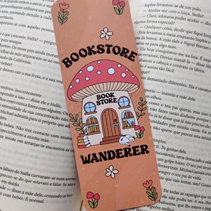 Könnte beinhalten: Ein skurriles Lesezeichen mit einem pilzförmigen Buchladen-Design. Das Lesezeichen ist pfirsichfarben mit den Worten "Bookstore Wanderer" und floralen Akzenten. Ein bezauberndes Accessoire.