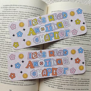 Pode incluir: Dois marcadores coloridos com o texto "Let's Read Another Chapter" em uma fonte divertida. Os marcadores apresentam flores, estrelas e rostos sorridentes, perfeitos para os amantes de livros. Os marcadores estão em cima de um livro aberto.