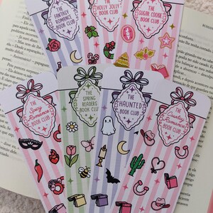 Könnte beinhalten: Ein Set aus sechs rechteckigen Lesezeichen aus Papier mit vertikalen Streifen in Pastellfarben. Jedes Lesezeichen zeigt oben ein dekoratives Etikett mit einer Schleife und Text wie The Fantasy Romance, Holly Jolly, Sugar Cookie, Spicy Romance, Spring Readers, The Haunted und The Cowboy Romance Book Club, ergänzt durch verschiedene kleine thematische Symbole.