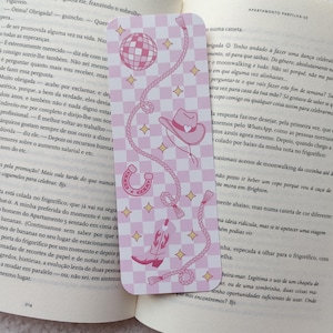Pode incluir: Marcador de livros rosa e branco com um design de bola de discoteca, chapéu de cowboy, ferradura e corda. O marcador de livros é retangular com cantos arredondados e um fundo xadrez.