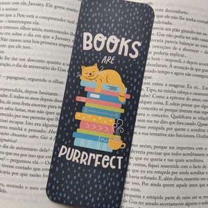 Può includere: Un segnalibro blu navy con le parole "BOOKS ARE PURRRFECT" in bianco. Un gatto giallo dorme su una pila di libri colorati. Una tazza di caffè gialla è accanto ai libri. Il segnalibro è lungo 15 cm.