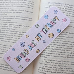 Pode incluir: Um marcador de página retangular com fundo roxo claro apresentando o texto BOOKS ARE MY THERAPY em uma fonte multicolorida, cercado por pequenas ilustrações de flores coloridas e ícones de rostos sorridentes amarelos e azuis.