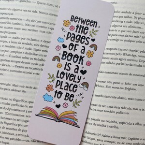 May include: White bookmark with the text "Between the pages of a book is a lovely place to be" in black, with colorful floral and rainbow accents. The bookmark is placed in an open book.