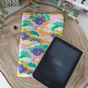 Pode incluir: Um leitor de e-books Kobo com uma tela preta e um contorno branco de um livro aberto. O leitor de e-books está sobre uma superfície de madeira ao lado de uma capa de tecido colorida com um padrão de nuvens e estrelas.