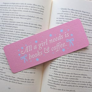 Può includere: Un segnalibro rettangolare in legno rosa con il testo azzurro "All a girl needs is books & coffee." Il design include piccole stelle azzurre e due piccoli fiocchi azzurri, posizionato sopra un libro aperto con testo stampato.