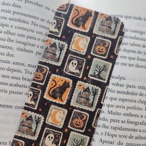 Pode incluir: Um marcador de livros com tema de Halloween com um padrão repetido de selos. Os selos apresentam ilustrações de gato preto, casa assombrada, abóbora, fantasma, lua e árvore. O marcador de livros é retangular com cantos arredondados.