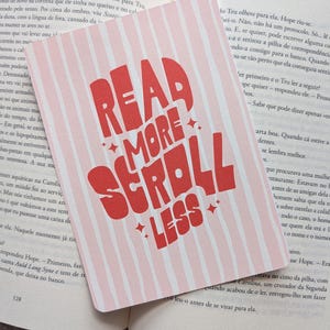 Könnte beinhalten: Ein rosa-weiß gestreifter Lesezeichen mit dem Text "READ MORE SCROLL LESS" in Rot. Das Lesezeichen ist rechteckig und liegt auf den offenen Seiten eines Buches.
