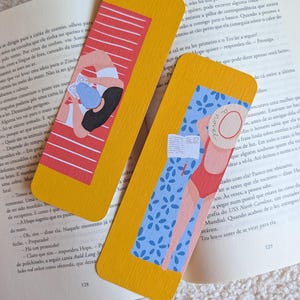 Pode incluir: Dois marcadores de página retangulares com cantos arredondados e bordas amarelas. Um mostra uma pessoa lendo em uma toalha listrada de vermelho e branco. O outro mostra uma pessoa de maiô vermelho e chapéu de palha lendo em uma toalha azul com estampa floral.