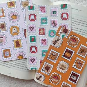 Puede incluir: Tres marcapáginas decorativos con un diseño de sello postal con ilustraciones temáticas de otoño e invierno. Los marcapáginas son rosas, crema y naranjas, con imágenes de calabazas, hojas y suéteres.