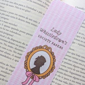 Pode incluir: Um marcador de livros rosa e branco xadrez com o texto "Lady Whistledown's Society Papers". O marcador de livros tem uma silhueta emoldurada em ouro e um laço rosa. O marcador de livros está colocado num livro aberto.