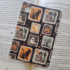 Op de afbeelding: Zwart-oranje Halloween bladwijzer met herhaalde postzegelontwerpen. Elke postzegel toont een zwarte kat, een spookhuis, een geest, een pompoen, een maan en een boom. De bladwijzer ligt op de pagina's van een boek.