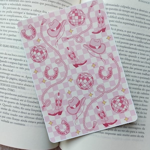 Pode incluir: Um cartão com padrão rosa e branco com chapéus de cowboy, botas, ferraduras, bolas de discoteca e desenhos de corda. O cartão está em um livro aberto.