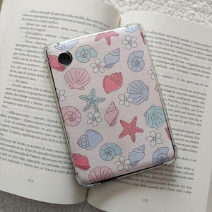 Può includere: Una custodia per telefono rosa con un motivo a conchiglie e stelle marine. La custodia è trasparente e mostra un libro con testo in portoghese.