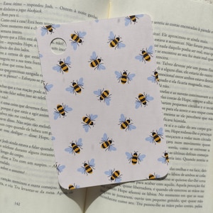 Könnte beinhalten: Rechteckiger Lesezeichen mit einem sich wiederholenden Muster aus gelben und schwarzen Bienen mit blauen Flügeln auf weißem Hintergrund. Das Lesezeichen liegt auf einem offenen Buch.
