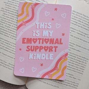 Pode incluir: Um cartão retangular rosa com o texto "THIS IS MY EMOTIONAL SUPPORT KINDLE" em branco e vermelho. O cartão apresenta um design ondulado com corações, estrelas e flores.