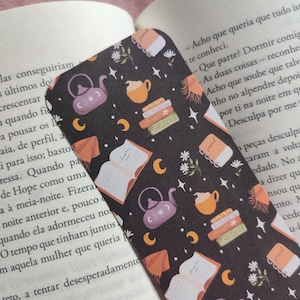 Pode incluir: Um marcador de livros preto com um padrão repetido de bules, livros, xícaras, luas e estrelas. O marcador de livros está em um livro aberto. O design inclui elementos laranja, roxos e brancos.