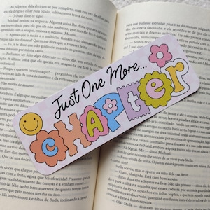 May include: A colorful bookmark with the text "Just One More Chapter" in a playful font. The bookmark features a smiley face and flower graphics, perfect for marking your place in a book.