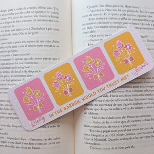 Pode incluir: Um marcador de livros retangular com quatro quadrados com desenhos florais em rosa e amarelo. O texto "Betty, IN THE GARDEN, WOULD YOU TRUST ME? - James" está impresso no marcador.