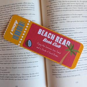 Pode incluir: Um marcador de página retangular com design de bilhete, com uma seção amarela à esquerda contendo um código de barras e o texto Admin 1, e uma seção vermelha à direita com o texto BEACH READ Book Club, Enjoy the Summer Sun, Sand and a Stack of Good Books.