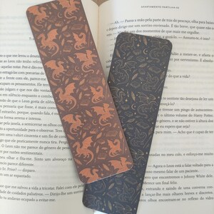 Pode incluir: Dois marcadores decorativos com um padrão de dragão. Um marcador é castanho escuro com dragões laranja, e o outro é azul escuro com um design semelhante. Os marcadores estão em cima de um livro aberto.