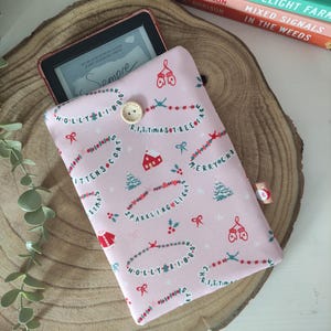 Può includere: Una custodia rosa per e-reader con un motivo natalizio festivo con testo, alberi di Natale e guanti. La custodia ha una chiusura a bottone e contiene un e-reader. La custodia è su una superficie di legno.