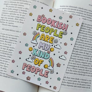 Può includere: Un segnalibro colorato con la scritta "BOOKISH PEOPLE ARE MY KIND OF PEOPLE" in un font giocoso, decorato con arcobaleni, fiori e stelle. Il segnalibro è inserito all'interno di un libro aperto.
