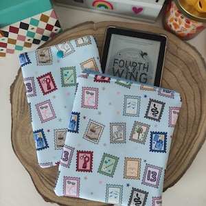 Puede incluir: Dos fundas de libro de tela azul y blanca con un patrón de sellos postales coloridos que representan diversas ilustraciones. Las fundas de libro están sobre una superficie de madera. Una funda de libro tiene el texto "FOURTH WING" en la funda.