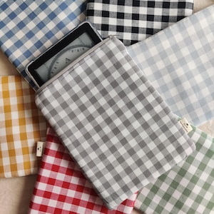 Könnte beinhalten: Eine Sammlung von Stoffhüllen in verschiedenen Farben, jeweils mit einem klassischen Vichy-Muster. Eine graue Hülle enthält einen E-Reader, dessen Bildschirm sichtbar ist. Die Hüllen sind in Blau-, Schwarz-, Grau-, Gelb-, Rot- und Grüntönen gehalten.