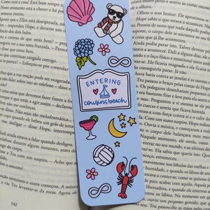 May include: Pale blue bookmark featuring illustrations of a seashell, a bear in sunglasses, flowers, and a lobster. The text "ENTERING cousins beach" is displayed on the bookmark.