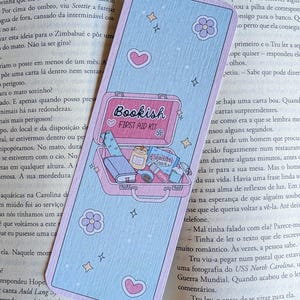 Könnte beinhalten: Ein rechteckiges Lesezeichen mit hellblauem Hintergrund und einer rosa Koffer-Illustration mit der Aufschrift Bookish First Aid Kit. Der Koffer enthält ein Buch, eine Kerze, einen Snack und Klebeband. Das Design zeigt kleine rosa Herzen, lila Blumen und gelbe Sterne.